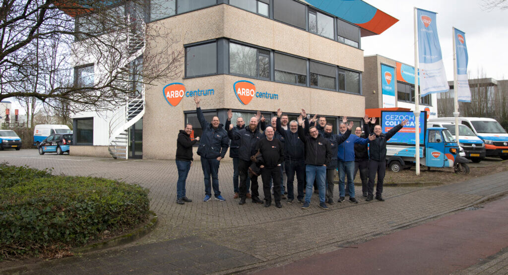 Voor het gebouw van het ARBOcentrum staat een groepje mensen, vrolijk de armen in de lucht stekend. De moderne faciliteit, met grote ramen en kleurrijke luifels, dient als knooppunt voor Tips voor het verbeteren van de veiligheidscultuur op de werkplek. In de buurt staan verschillende merkvoertuigen geparkeerd.
