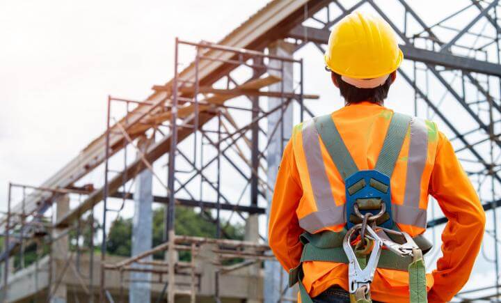 Een bouwvakker met een oranje veiligheidsvest, gele helm en harnas kijkt naar een stalen constructie met een steiger op een bouwplaats. De lucht is bewolkt op de achtergrond.