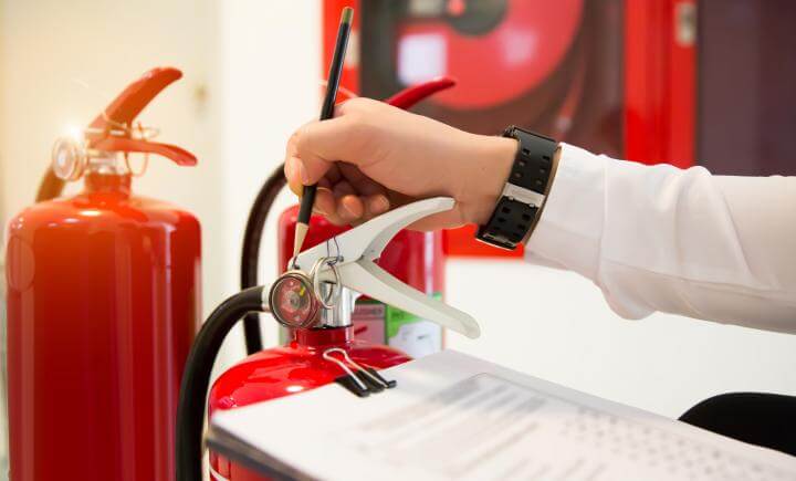 Een persoon in een wit shirt controleert de drukmeter van een rode brandblusser met een pen, en oefent brandpreventie. Een klembord met een checklist staat op de voorgrond, terwijl een andere blusser en veiligheidsuitrusting op de achtergrond zichtbaar zijn, klaar voor nieuwe handen om de voortdurende veiligheid te waarborgen.