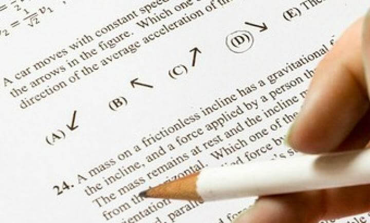 Een hand die een potlood vasthoudt, zweeft boven een natuurkundetoets. De toets bevat diagrammen met pijlen en een meerkeuzevraag over een massa op een wrijvingsloze helling, met opties (A), (B), (C) en (D).