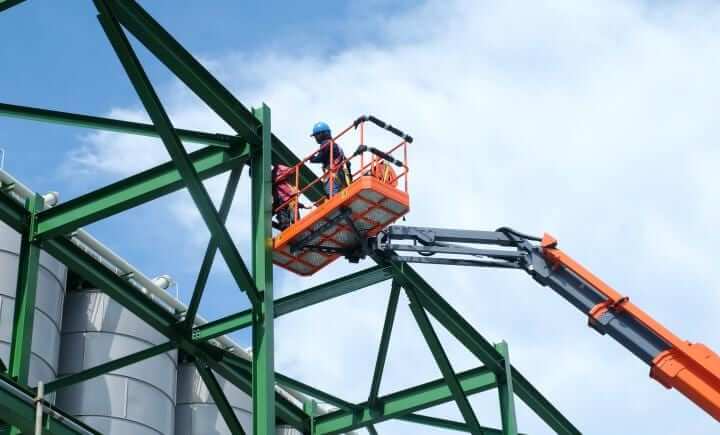 Twee arbeiders met helmen op staan op een oranje hydraulische lift, hoog gepositioneerd bij een groen stalen frame van een constructie. Ze voeren bouwwerkzaamheden uit onder een gedeeltelijk bewolkte lucht.