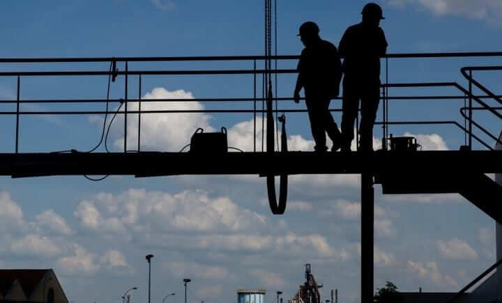 Silhouetten van twee bouwvakkers die op een verhoogd platform met een reling lopen tegen een achtergrond van een blauwe lucht met verspreide wolken. Industrieel materieel is in de verte zichtbaar.