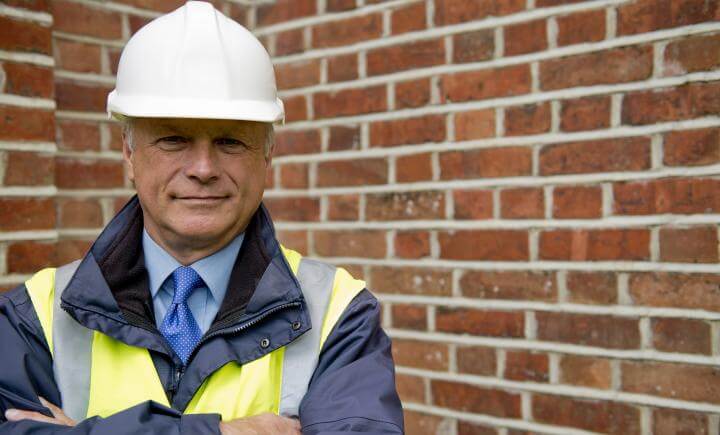 Een volwassen man met een witte helm, een reflecterend veiligheidsvest en een blauwe stropdas staat met gekruiste armen voor een bakstenen muur. Hij is buiten en lijkt zelfverzekerd.