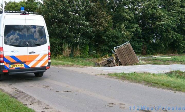Een politiebusje staat geparkeerd op een landweg naast een ingestorte houten constructie. Bomen en gras zijn zichtbaar op de achtergrond. De lucht is gedeeltelijk bewolkt en er liggen bandensporen op de weg.