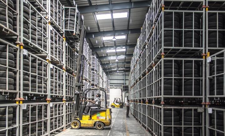 Een magazijn vol met grote stapels banden opgeslagen op metalen rekken. Twee gele vorkheftrucks zijn zichtbaar; één rijdt tussen de rijen en de andere staat verderop in het gangpad geparkeerd. Het plafond heeft dakramen, waardoor er natuurlijk licht binnenkomt.