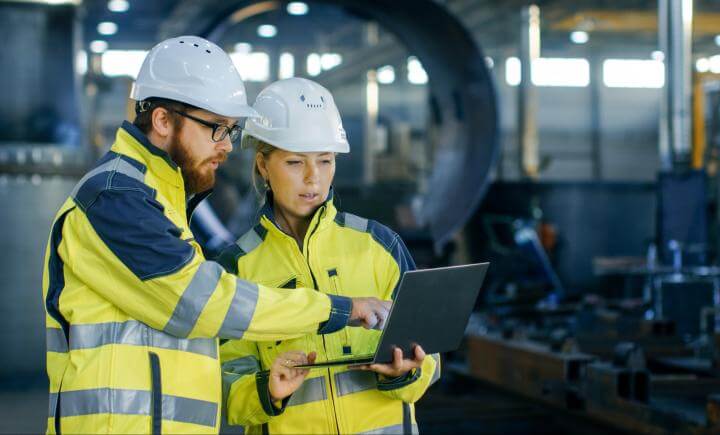 Twee personen in veiligheidsuitrusting, waaronder helmen en fluorescerende hesjes, bespreken een VCA-certificeringschecklist via een laptop in een industriële omgeving met machines op de achtergrond. De scène is goed verlicht en professioneel.