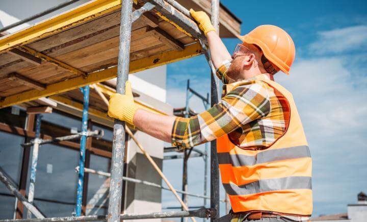 Een bouwvakker met een oranje veiligheidsvest, helm en handschoenen is bezig met het verstellen van steigers op een bouwplaats. De lucht is helder en blauw, wat duidt op een zonnige dag. Gefocust op het vastzetten van de metalen constructie, laat hij een van de 3 situaties zien waarbij je een rolsteiger gebruikt.