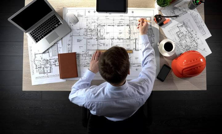 Een persoon werkt aan een bureau met architectonische blauwdrukken, een laptop en een koffiekopje. Een oranje helm ligt in de buurt terwijl ze nauwkeurig op de plannen tekenen met een liniaal en potlood, om ervoor te zorgen dat elk detail overeenkomt met de RI&E-normen.