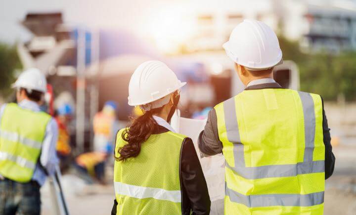 Twee bouwvakkers in fluorescerende vesten en helmen bekijken VIL-VCU-examenblauwdrukken op een bouwplaats. Ze staan met hun rug naar de camera en concentreren zich op het lopende werk op de achtergrond, waar wazige werknemers en apparatuur zichtbaar zijn.