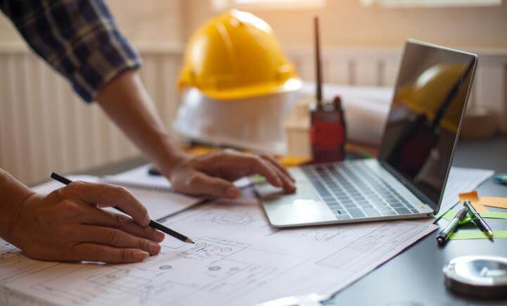 Een persoon die aan een bureau werkt met architectonische blauwdrukken, met behulp van een laptop en een potlood. Een gele helm, een portofoon en verschillende kantoorbenodigdheden zijn zichtbaar. Zonlicht stroomt door een raam, wat een warme sfeer creëert.