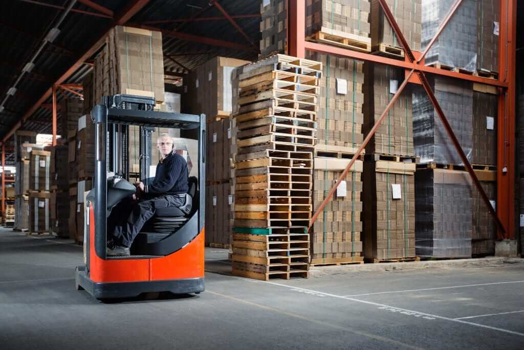 Een persoon bedient een vorkheftruck in een groot magazijn vol met stapels dozen en pallets, en toont trots zijn Reachtruck-certificaat. Het magazijn heeft hoge, rode metalen balken en rekken vol met kartonnen dozen, terwijl de vloer is gemarkeerd met geverfde lijnen.