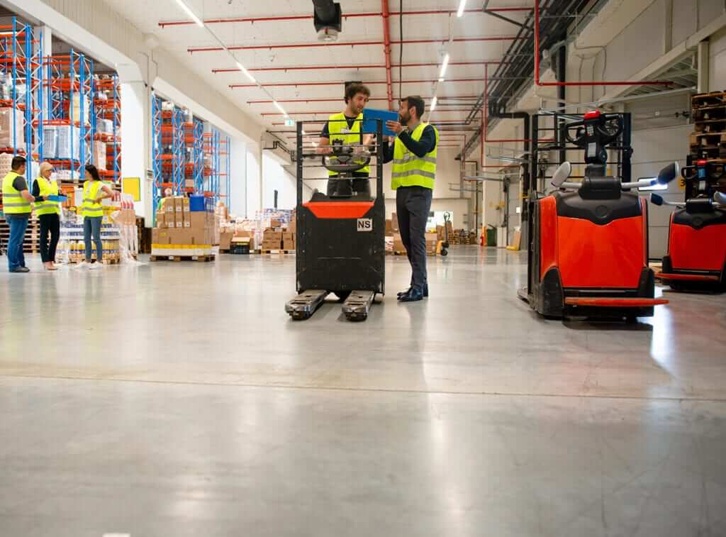 Werknemers in een magazijn, gesponsord en gekleed in gele veiligheidsvesten, inspecteren documenten op een vorkheftruck. Op de achtergrond beheren anderen pallets en inventaris. Twee elektrische palletverplaatsers staan in de buurt op de gladde betonnen vloer.