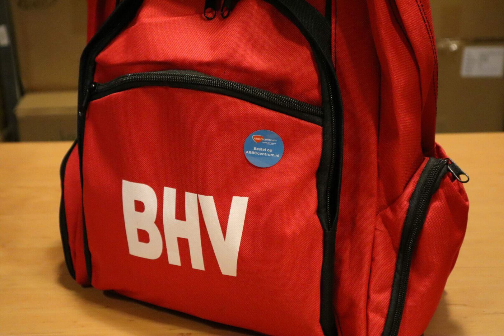 Een rode rugzak met grote witte letters BHV op de voorkant staat op een houten oppervlak. Ideaal voor een EHBOer bij evenementen, met meerdere ritsvakken en een ronde blauwe knop met tekst.
