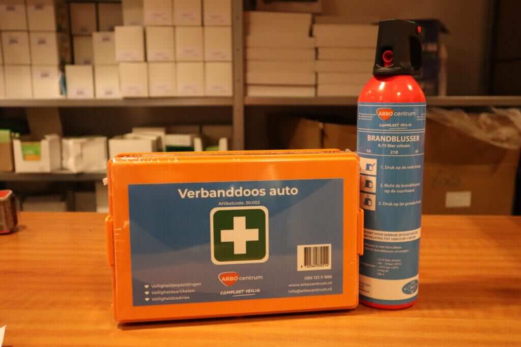 Op een houten tafel staan een oranje EHBO-doos voor in de auto en een rode brandblusser uitgestald, wat de essentie van veilig met de auto op vakantie belichaamt. Op de achtergrond staan planken met diverse dozen, wat doet denken aan een opslagruimte of voorraadkamer.