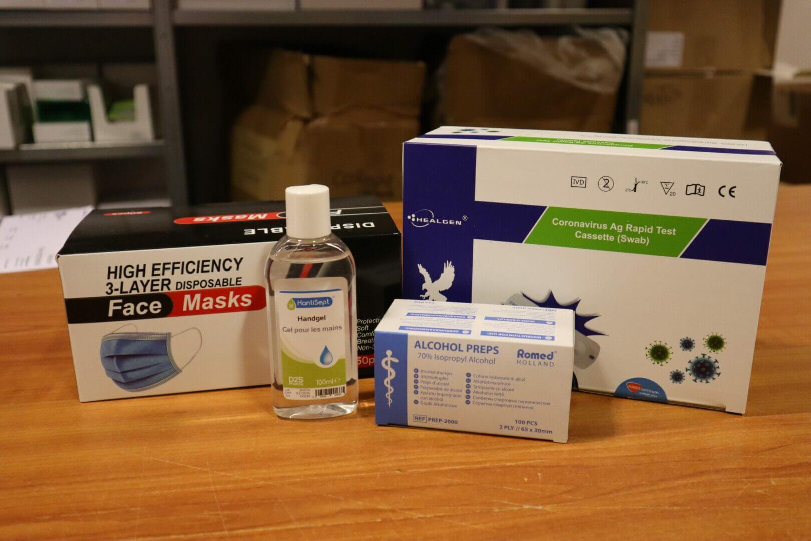 Gezondheidszorgbenodigdheden op een tafel, met een doos met wegwerpmaskers, handontsmettingsmiddel, alcoholpreppads en corona-sneltestcassettes: essentieel om veilig op vakantie te blijven tijdens een pandemie. Op de achtergrond zijn schappen gevuld met extra benodigdheden.