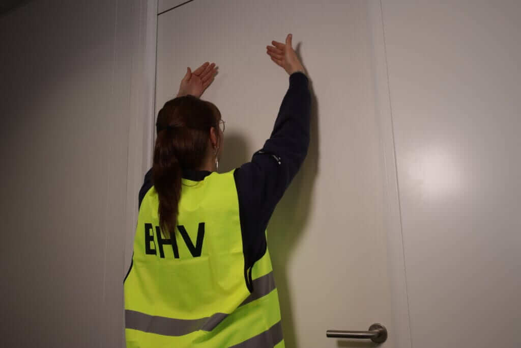 Een persoon die een felgeel vest draagt met BHV op de rug, reikt naar een gesloten deur. De persoon heeft lang haar dat naar achteren is gebonden en staat binnen. De kamer heeft witte muren en een moderne deurknop.