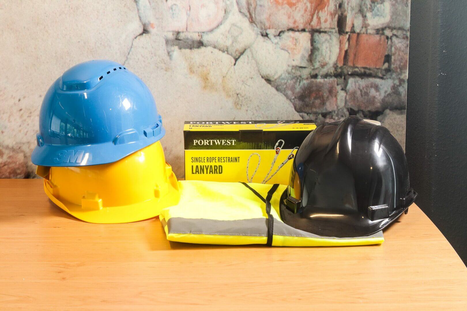 Een houten tafel toont twee bouwhelmen, een blauwe en een zwarte, naast een geel en grijs veiligheidsvest en een doos met het label Portwest Single Rope Restraint Lanyard, essentiële benodigdheden voor degenen die de VCA-cursus afronden. Een getextureerde muur dient als achtergrond.