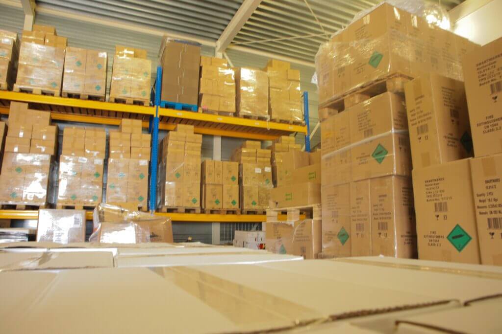 Een magazijn vol met stapels kartonnen dozen op metalen planken en pallets. De dozen zijn verpakt in plastic en de setting is goed verlicht, met hoge plafonds en een nette indeling.