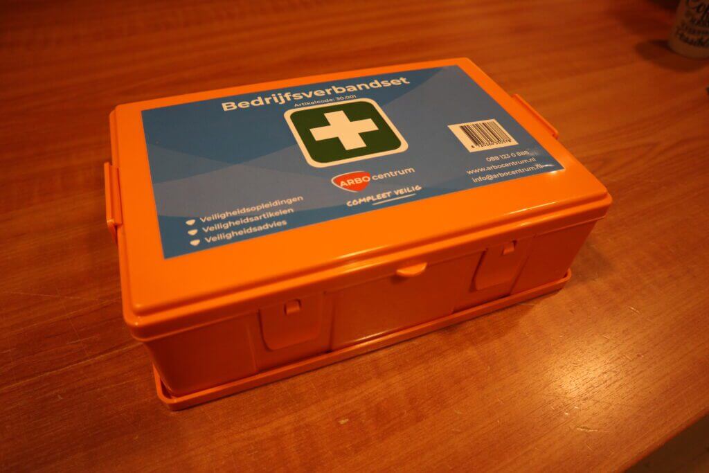 Een oranje EHBO-doos met het opschrift Bedrijfsverbandset staat op een houten oppervlak. Het deksel, versierd met een groen kruissymbool en contactgegevens, lijkt klaar om verschillende noodgevallen aan te pakken. Het zet aan tot nadenken: Welke vormen van epilepsie zijn er?.