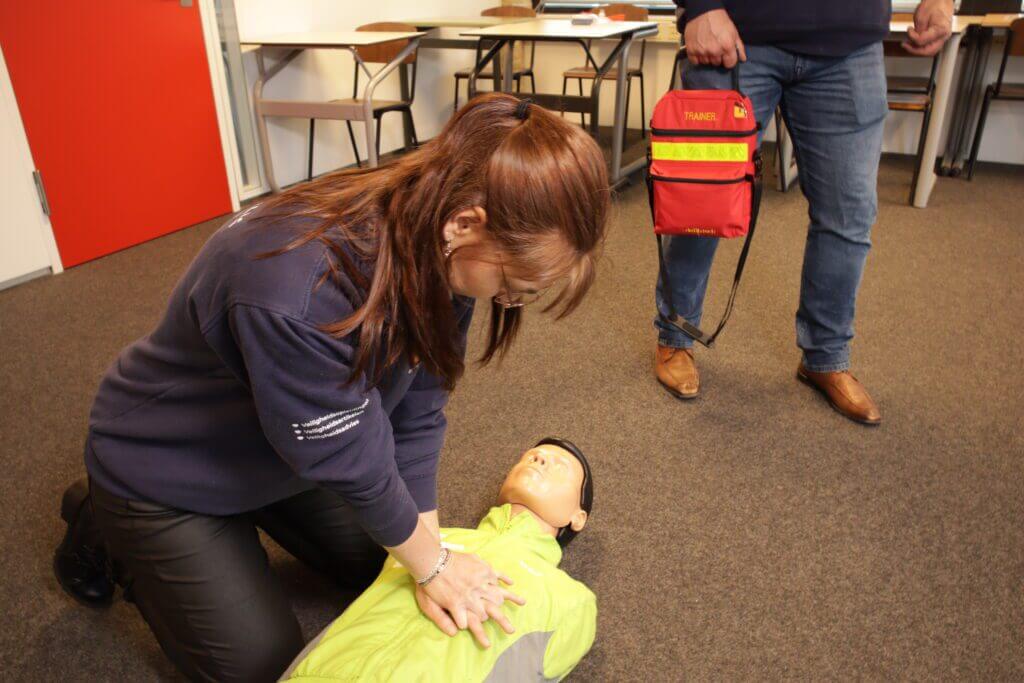 In een klaslokaal knielt iemand op de grond en voert borstcompressies uit op een reanimatiepop, terwijl een ander in de buurt staat met een rode EHBO-doos. De scène weerspiegelt een belangrijke les: hoe herken je een hartaanval? Stoelen en tafels vullen de achtergrond.