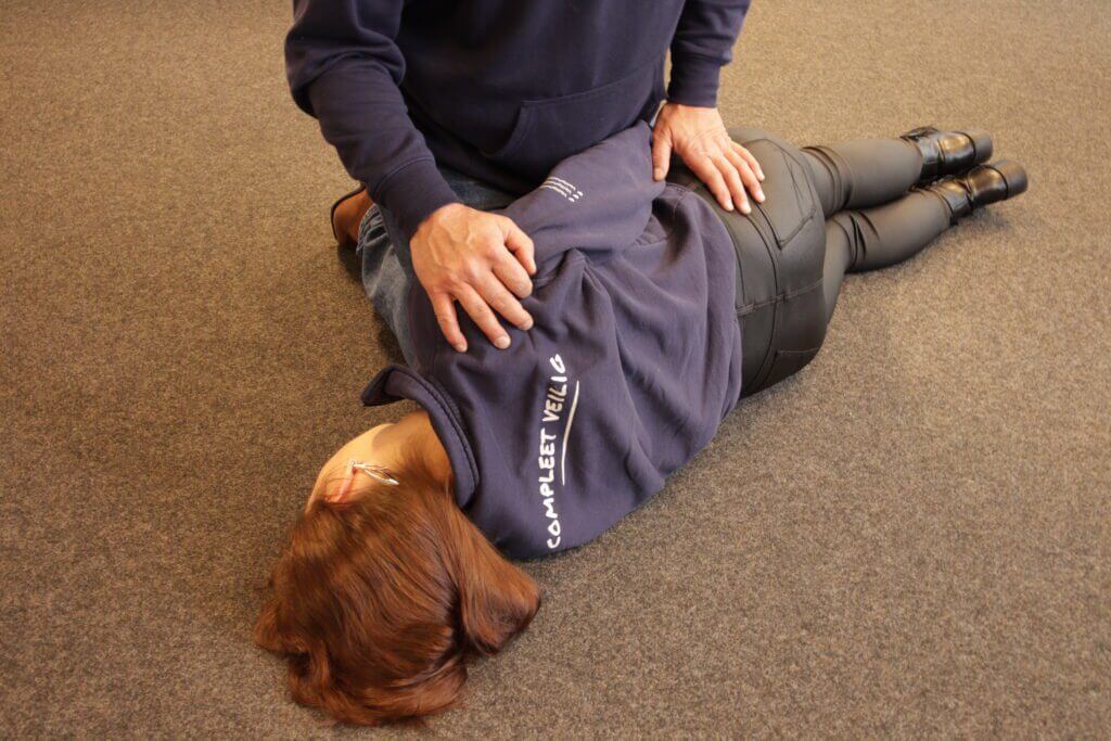 Een persoon in een blauwe trui en zwarte broek ligt op zijn zij, terwijl een ander naast hem knielt en een hand op zijn schouder en heup legt. Deze scène zou de vraag kunnen beantwoorden: Waarom stabiele zijligging?, aangezien het mogelijk een herstelhoudingstechniek op een vloer met tapijt laat zien.