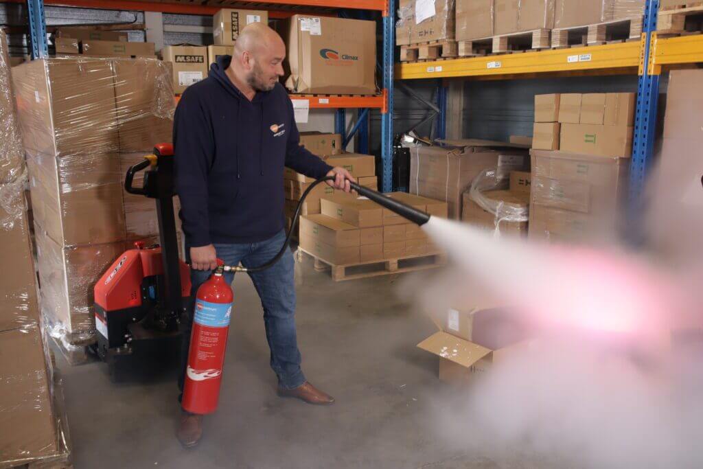 In een magazijn vol met planken en opgeslagen artikelen demonstreert een persoon het gebruik van een brandblusser, waarbij hij een wolk witte rook verspreidt over een kartonnen doos. Deze scène onderstreept dat veiligheid verder gaat dan producten, en benadrukt het belang van veiligheidspraktijken die verder gaan dan alleen goederen.