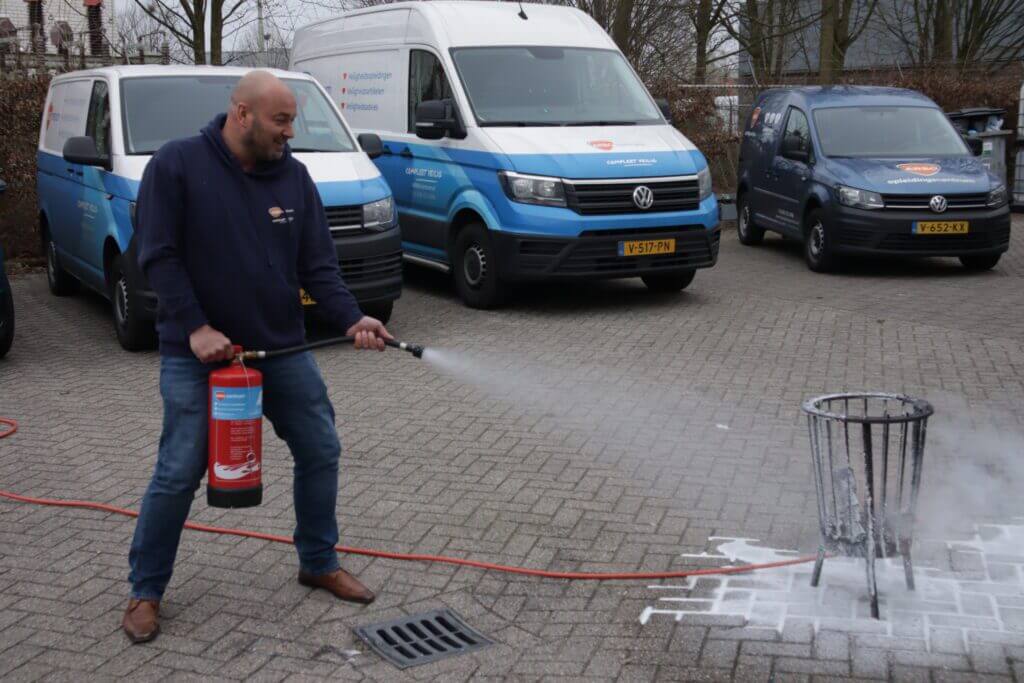 Een man gebruikt snel een brandblusser op een brandende afvalbak buiten, waarmee hij een van de 5 redenen om meer BHV’ers binnen jouw bedrijf te hebben laat zien. Verschillende geparkeerde busjes vormen het decor in dit bruisende parkeertafereel.