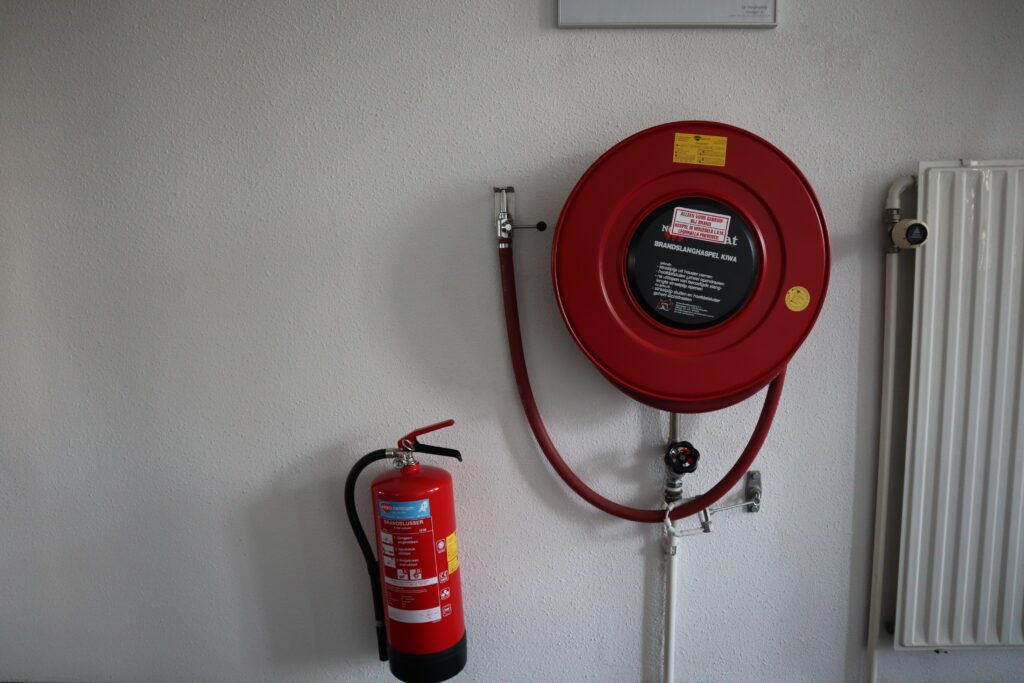 Een rode brandslanghaspel gemonteerd op een witte muur met daaronder een brandblusser benadrukt de veiligheidsvoorbereiding. Aan de rechterkant is een radiator zichtbaar, wat drie redenen onderstreept om een beheerder brandmeldinstallatie cursus te volgen: uitgebreide kennis, naleving en efficiƫnte respons.
