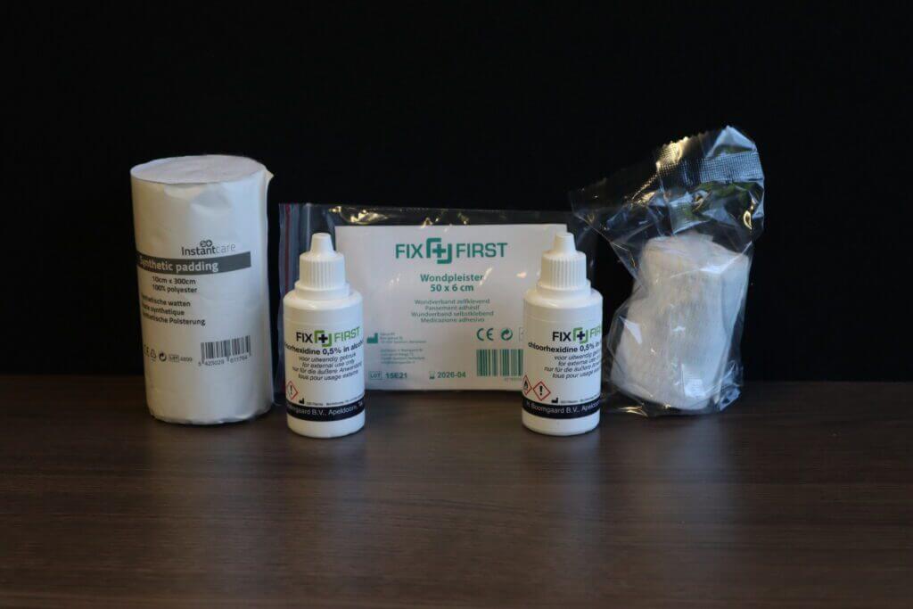 Op een slecht verlichte tafel ligt een verzameling medische benodigdheden, waaronder een rol wattenstaafjes, twee flessen met het opschrift FIX FIRST, wat de redenen om desinfectiemiddelen te gebruiken benadrukt, naast wondverbanden en een zakje gaas.