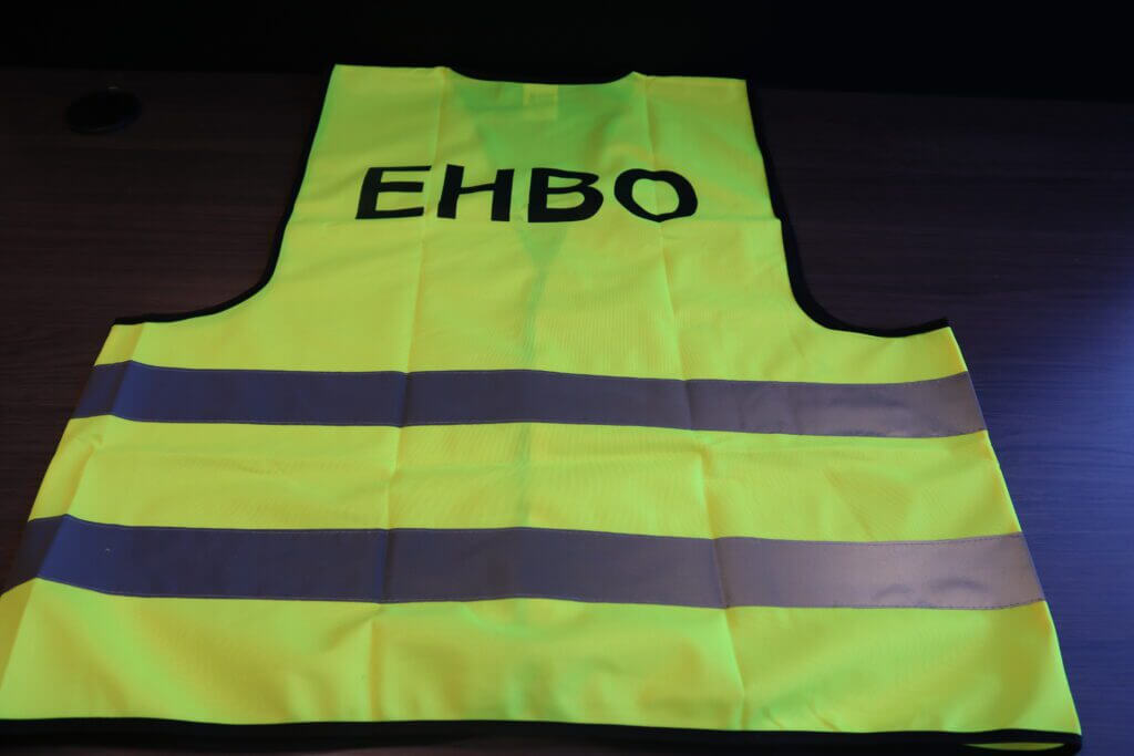 Een geel veiligheidsvest met reflecterende grijze strepen ligt op een donker oppervlak, met de dikke zwarte letters EHBO op de achterkant. Het is duidelijk waarom een EHBO-hesje aan te raden is voor zichtbaarheid en veiligheid.