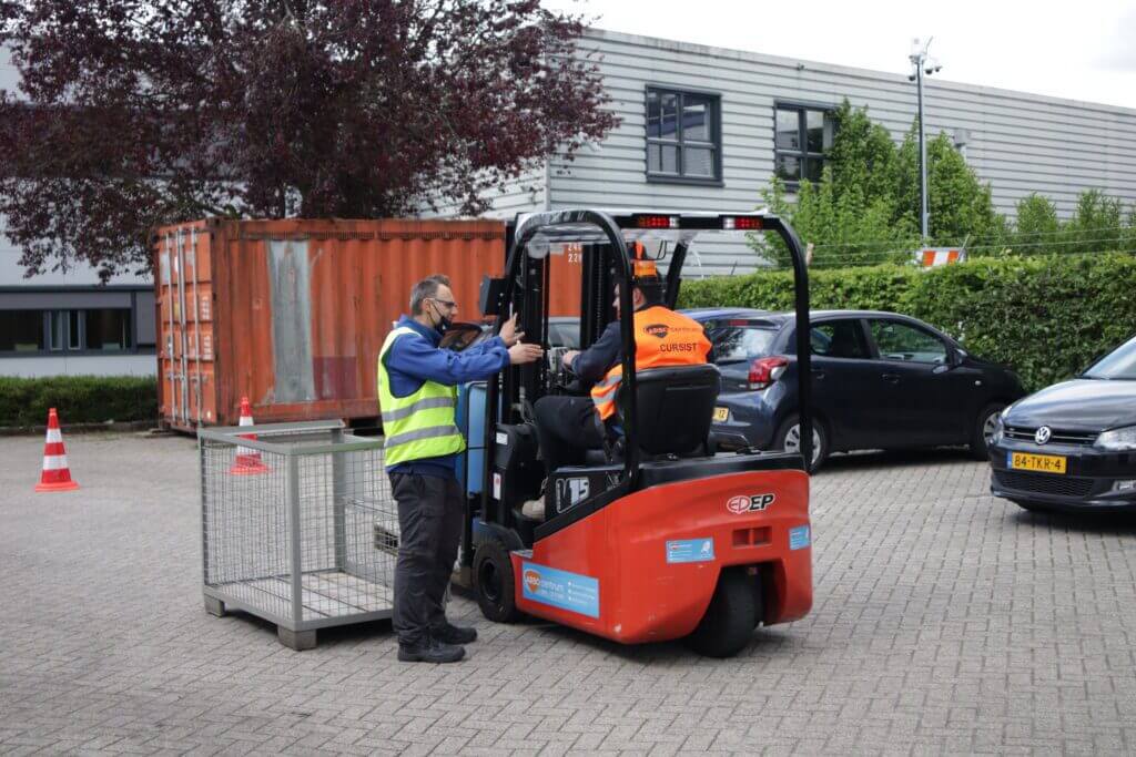 Twee mensen zijn buiten bezig met een heftrucktraining. De instructeur in een geel vest gebaart naar de heftruckchauffeur in een oranje vest, met de nadruk op de gevolgen van het rijden zonder heftruckcertificaat. Een metalen krat is aan de heftruck bevestigd, met een grote oranje container en verschillende autos op de achtergrond.