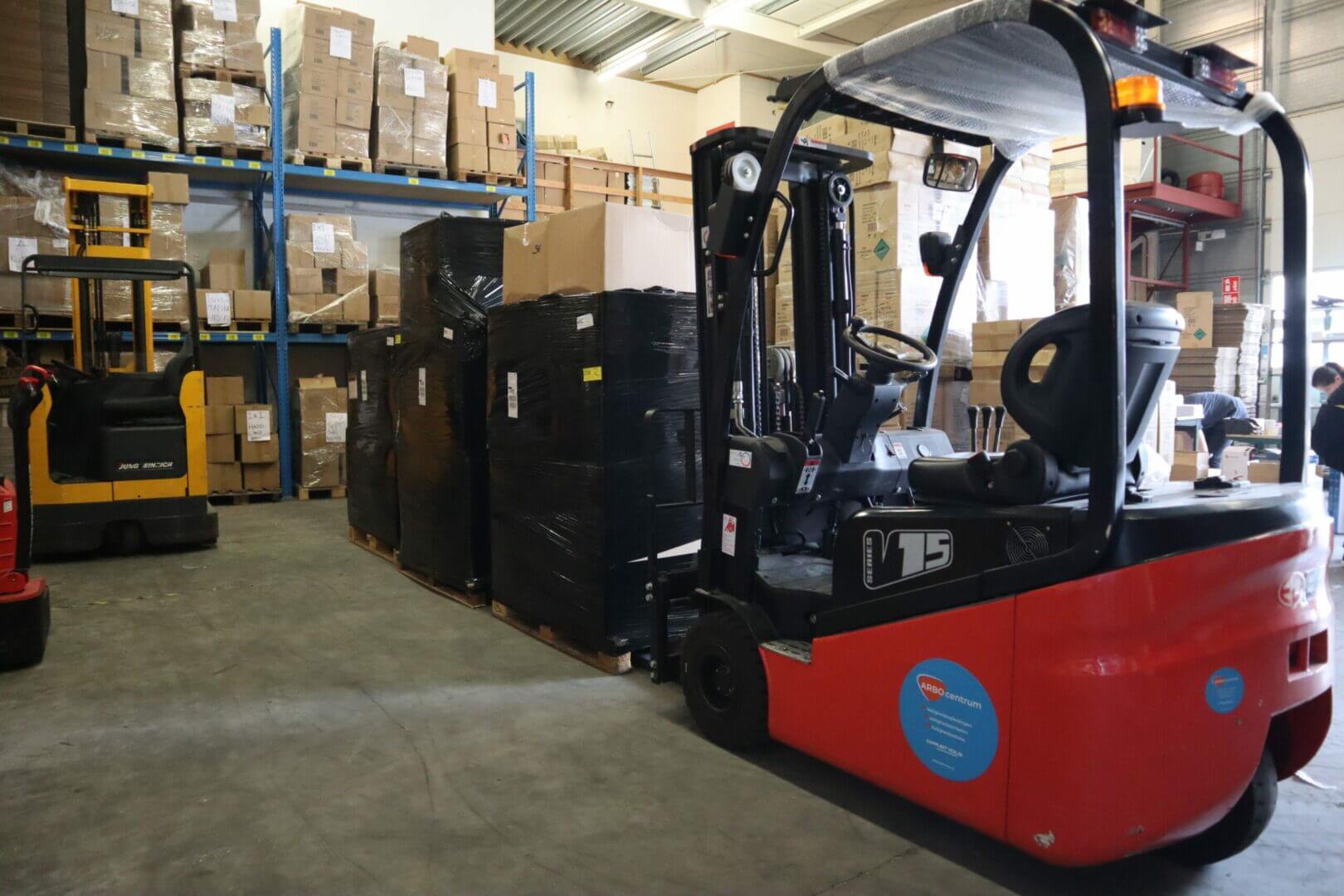 Een rode vorkheftruck vervoert meerdere pallets verpakt in zwart plastic in een magazijn, wat benadrukt waarom aparte certificeringen voor vorkheftrucks en reachtrucks cruciaal zijn. Planken met kartonnen dozen staan op de achtergrond, een andere vorkheftruck staat in de buurt geparkeerd en rechts is een persoon gedeeltelijk zichtbaar.