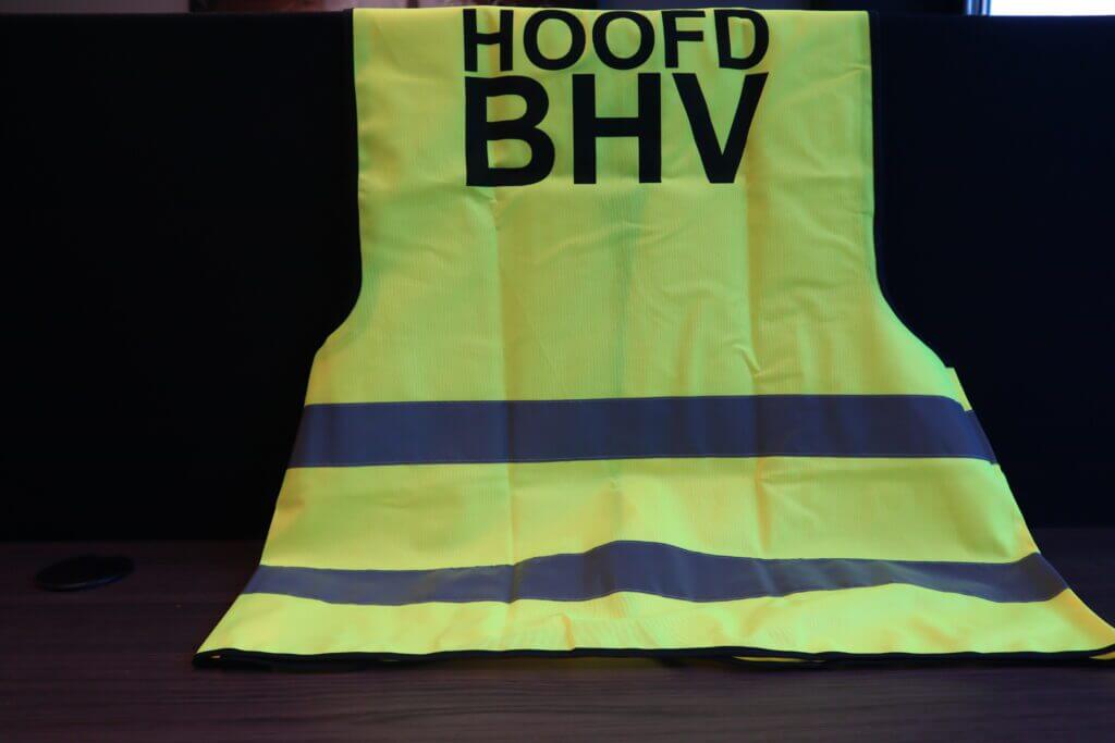 Een fluorescerend geel veiligheidsvest wordt op een donker oppervlak getoond. Het vest, gemerkt met HOOFD BHV, benadrukt het verschil tussen hoofd BHV en ploegleider met zijn kenmerkende ontwerp, met twee horizontale reflecterende strepen over de borst- en taillegebieden.