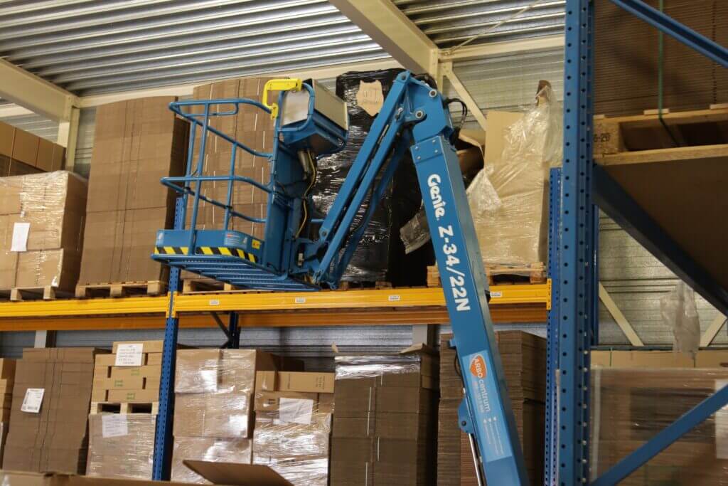Een bekwame hoogwerkerchauffeur bedient de blauwe Genie Z-34/22N lift en bereikt een hoge plank in het magazijn. Omringd door stapels kartonnen dozen en pallets op metalen planken, wordt het tafereel omlijst door metalen plafondbalken en een gele veiligheidsleuning op het platform van de lift.
