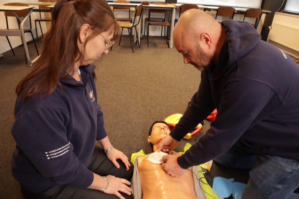 Twee mensen oefenen reanimatie op een pop, een essentiƫle vaardigheid die valt onder levensreddend handelen. De een drukt de borstkas samen terwijl de ander aandachtig toekijkt. Ze bevinden zich in een kamer met stoelen en een raam, toegewijd aan het onder de knie krijgen van deze levensreddende procedure.
