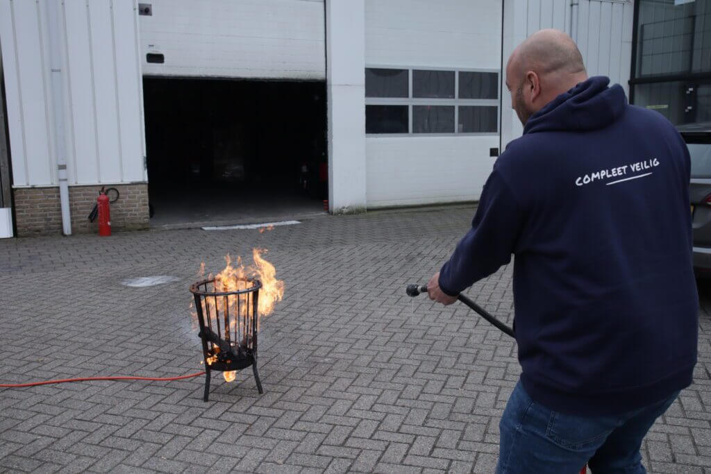 Een persoon in een blauwe hoodie demonstreert een van de 5 manieren om een kleine brand te bestrijden door een brandblusser te gebruiken op een kleine brand in een metalen mand buiten. Een andere brandblusser is zichtbaar bij de grote garage-ingang.
