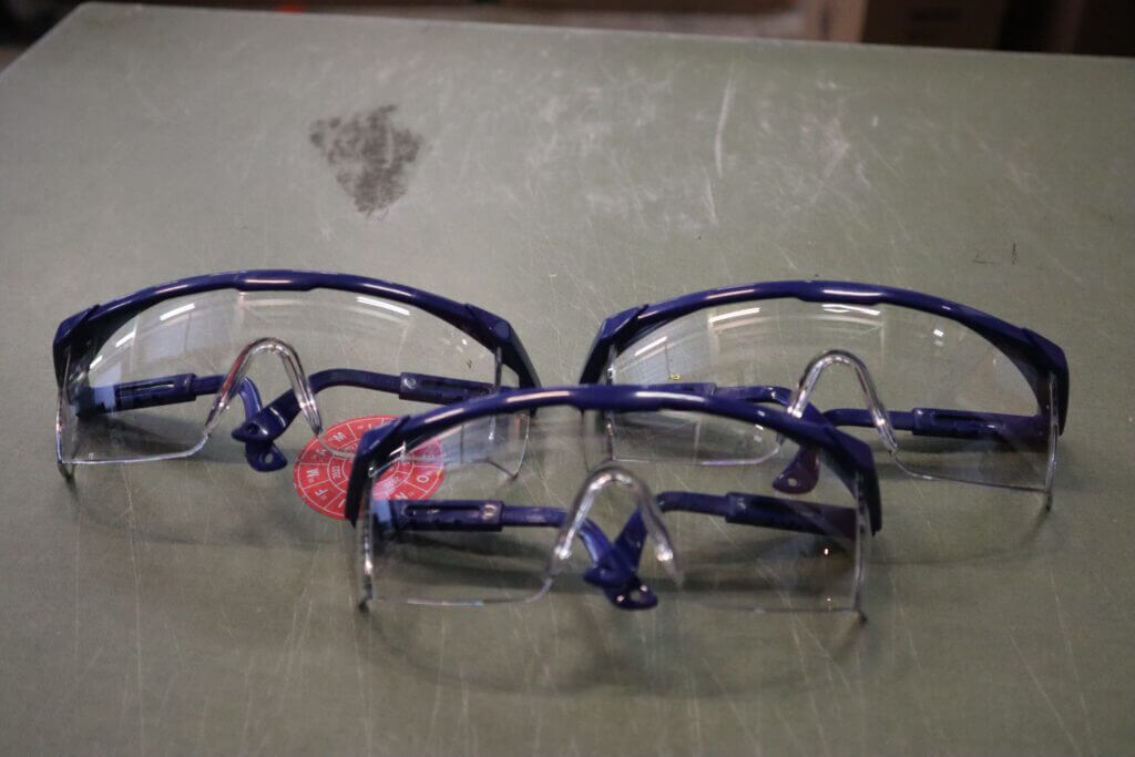 Drie paar veiligheidsbrillen met blauwe monturen en heldere lenzen zijn gerangschikt op een groen oppervlak. Een ronde rode sticker is gedeeltelijk zichtbaar onder een paar, wat het belang van nog veiliger werken met een veiligheidsbril op sterkte voor verbeterde bescherming benadrukt.