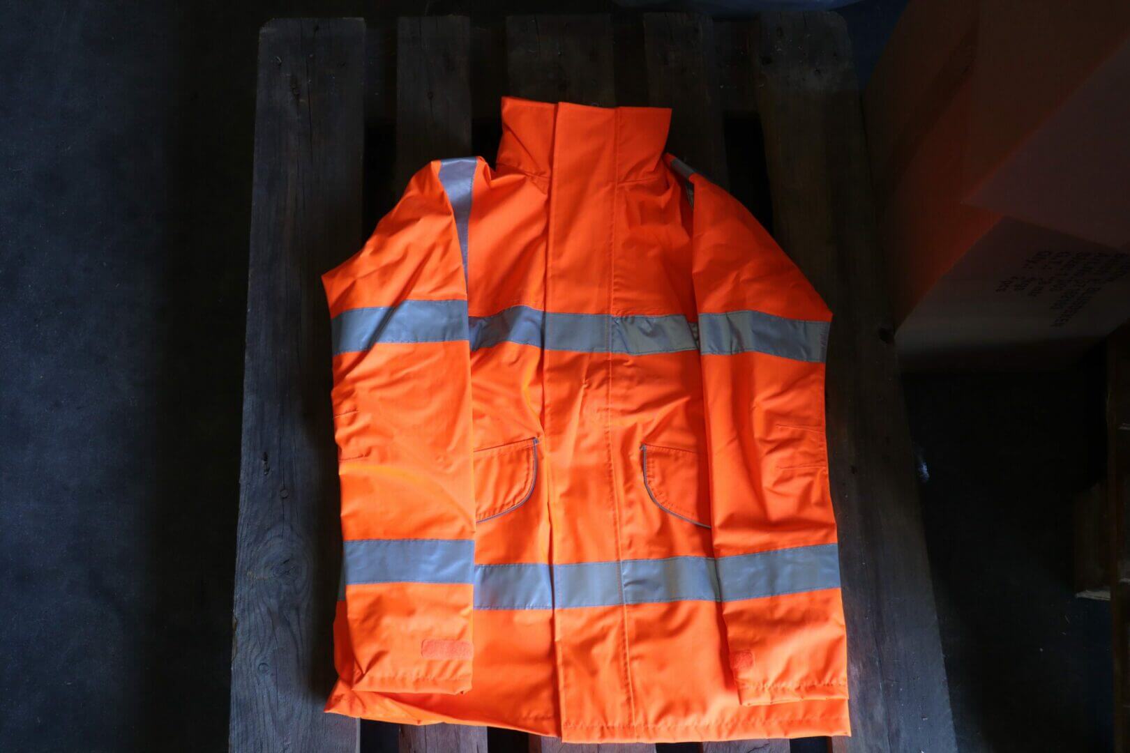 Een feloranje veiligheidsvest met reflecterende zilveren strepen ligt plat op een houten pallet, klaar voor die Aan de slag in veiligheidskleding-momenten. Ontworpen voor hoge zichtbaarheid, heeft het twee grote zakken aan de voorkant. De zwak verlichte achtergrond contrasteert met de felle kleur van het vest.