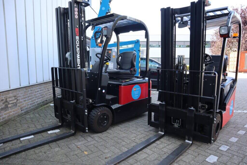 Twee zwarte vorkheftrucks staan geparkeerd op een geplaveid terrein bij een gebouw, hun vorken omhoog en de stoelen zichtbaar, wat echte voorbeelden illustreert voor degenen die tips zoeken om snel een heftruckchauffeur te worden. Een blauw stuk machinerie is gedeeltelijk zichtbaar op de achtergrond.