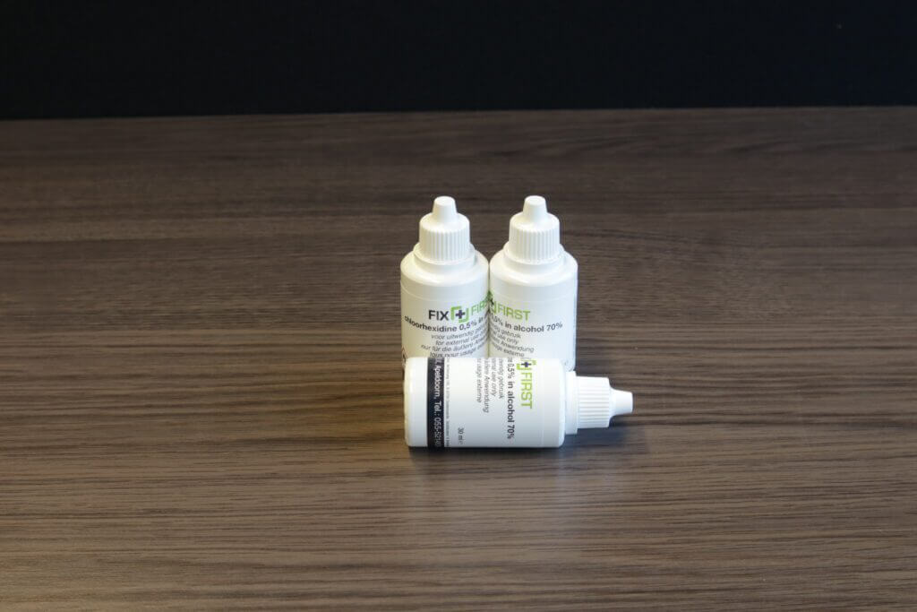 Drie kleine witte flesjes met druppeldopjes staan op een houten oppervlak tegen een donkere achtergrond. De etiketten, gedeeltelijk zichtbaar, bevatten tekst en een groen kruisontwerp dat verwijst naar de vraag: Wat is desinfectie?.