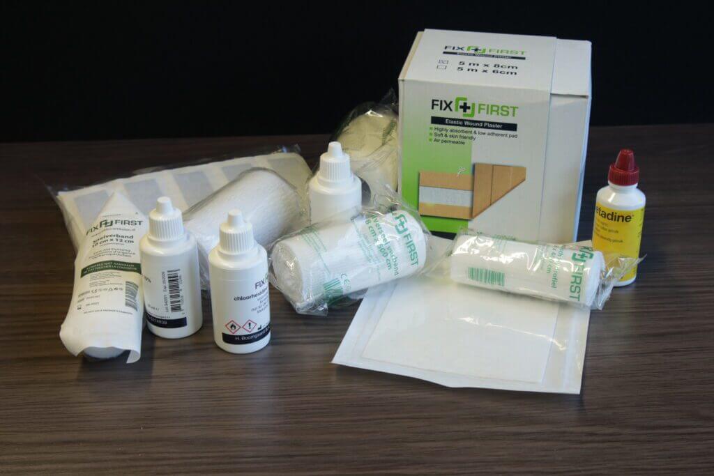 Op een houten oppervlak ligt een verzameling medische benodigdheden netjes uitgestald, met onder andere verbanden, flessen met antiseptische middelen, verbandsets en een doos met het opschrift FIX FIRST. Hierin staan tips voor het behandelen van een schaafwond, met essentiƫle instructies voor de basisverzorging van wonden.