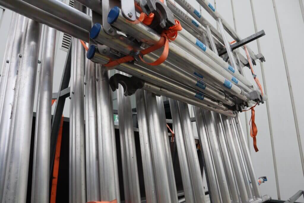 Een stapel gevouwen metalen ladders met oranje banden leunt tegen een lichtgekleurde muur, net als de veiligheidsmaatregelen die u aanzetten om ervoor te zorgen dat uw werknemers een rolsteigercertificaat krijgen. Elke ladder heeft blauwe eindkappen en het tafereel speelt zich binnen af.