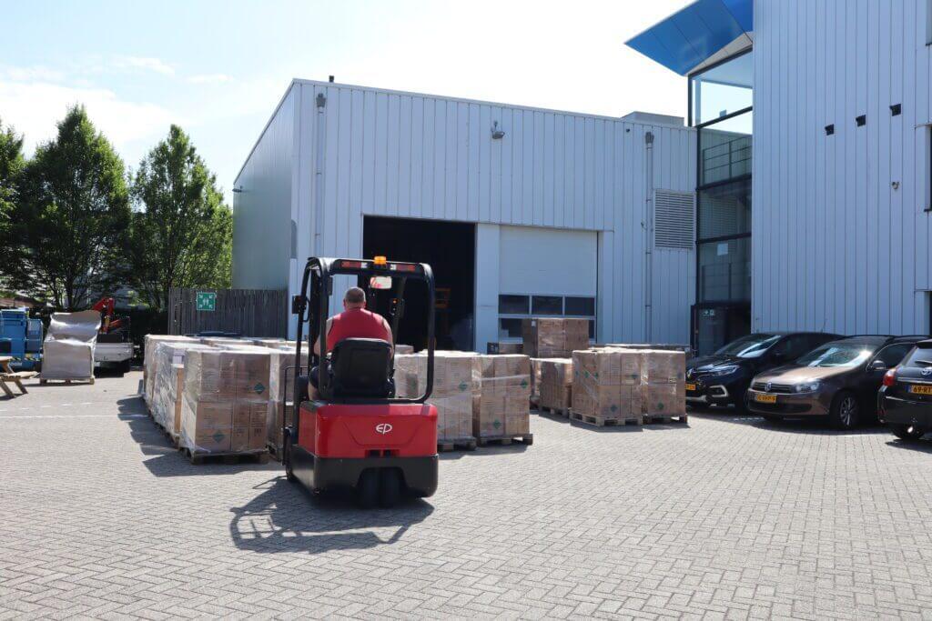 Een persoon op een rode heftruck verplaatst pallets met goederen buiten een groot industrieel magazijn, wat een van de 3 redenen om medewerkers VCA te laten halen benadrukt. Het gebouw is wit met een gedeeltelijk open deur. Meerdere autos staan rechts geparkeerd en bomen zijn zichtbaar op de achtergrond.