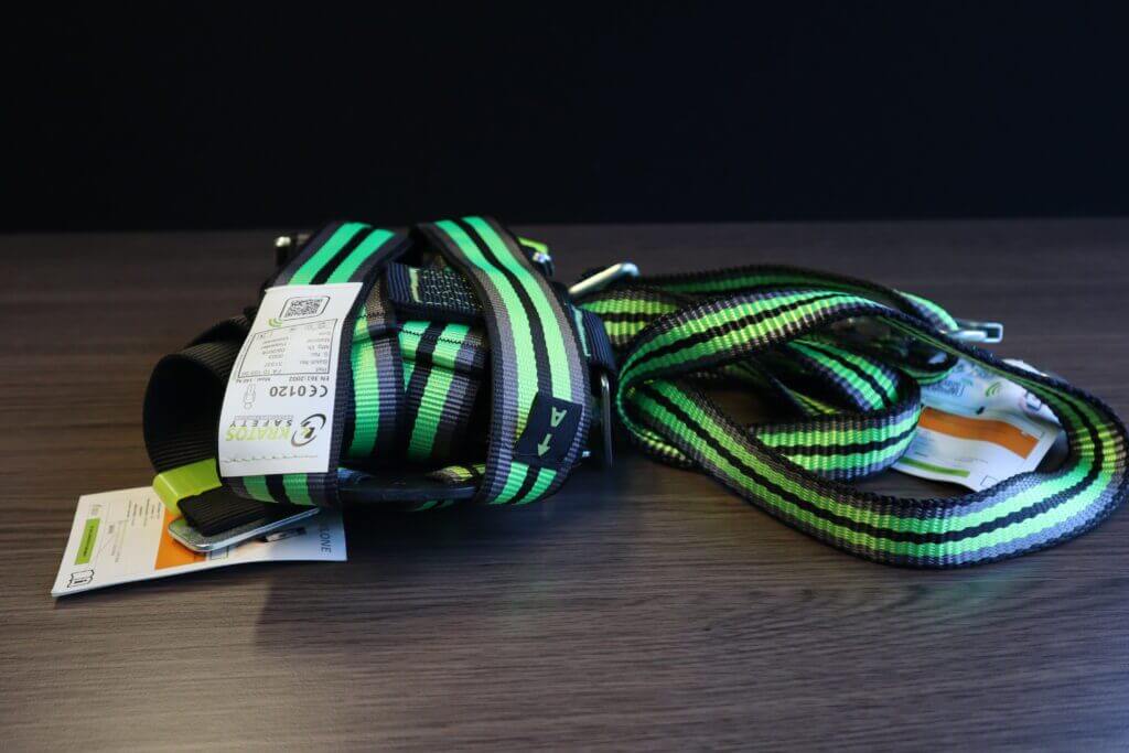 Een groen en zwart harnas met bevestigde labels ligt op een houten oppervlak, wat illustreert waarom het belangrijk is om met valbeveiliging te werken. Met meerdere verstelbare banden en metalen gespen, is het netjes opgerold naast andere essentiƫle uitrusting op tafel.