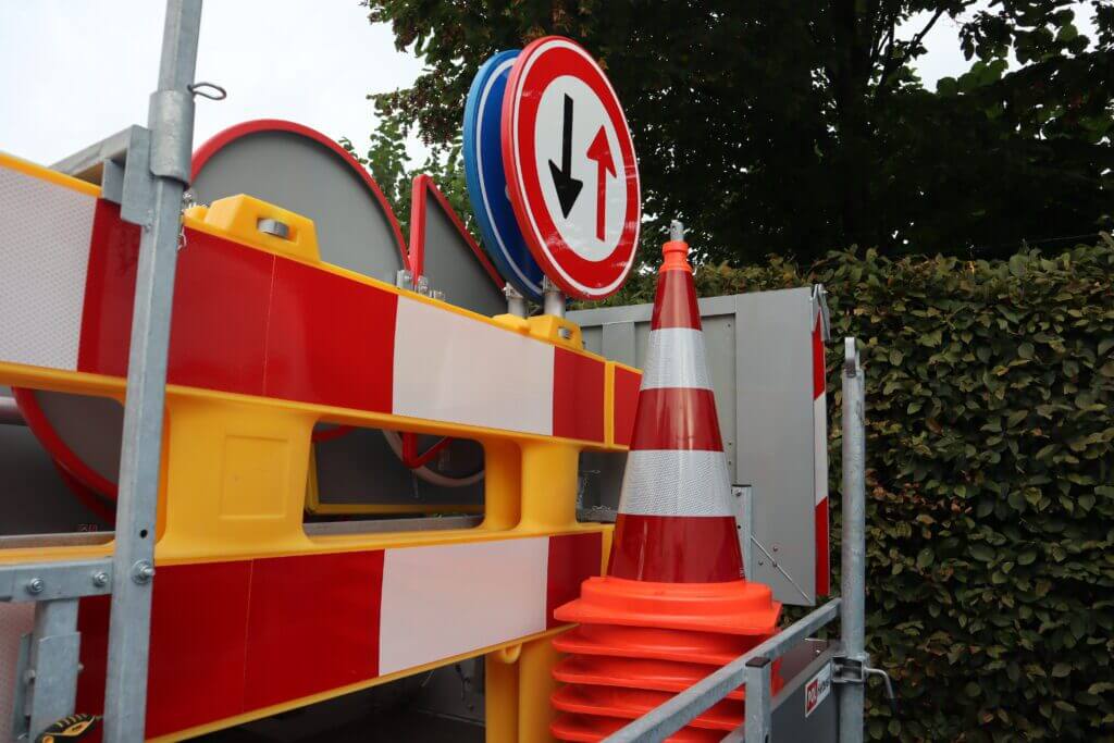 Verkeersapparatuur, die de 3 risicos van werken langs de weg benadrukt, omvat oranje kegels, rood-witte gevarenborden en een richtingaanwijzerbord bij een metalen hek. Struiken en bomen creëren een natuurlijke achtergrond.