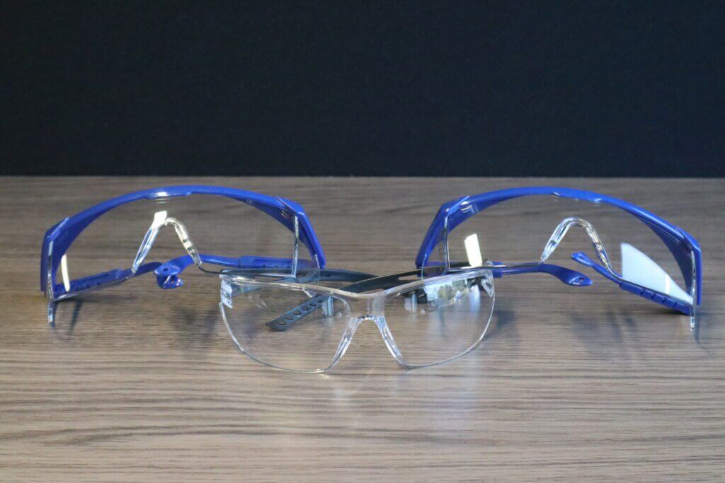 Drie paar veiligheidsbrillen op een houten oppervlak: twee met een blauw montuur en heldere glazen aan de achterkant, en één met een helder montuur en heldere glazen aan de voorkant. Dit is waarom je altijd een veiligheidsbril moet dragen—een zwarte achtergrond versterkt hun prominentie.