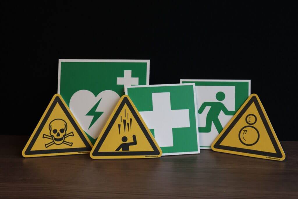 Een verzameling veiligheidsborden op een houten oppervlak tegen een zwarte achtergrond illustreert redenen om veiligheidspictogrammen binnen uw bedrijf te hangen. De borden bevatten een doodshoofd met gekruiste beenderen, een figuur die onder puin valt en meer, die allemaal cruciaal bewustzijn op de werkplek promoten.