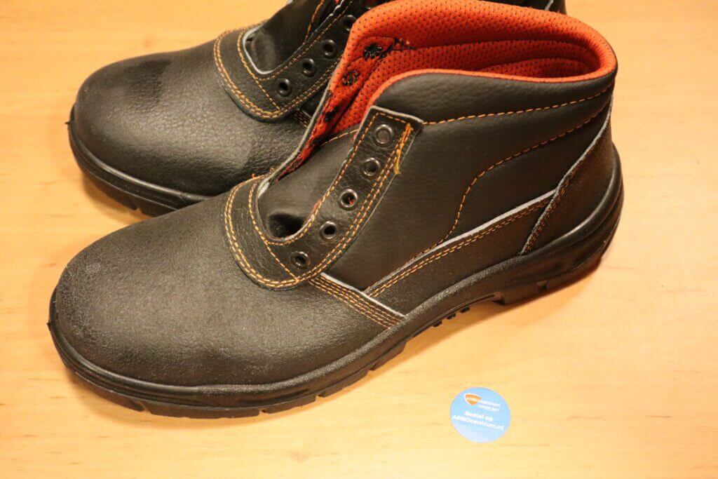 Een paar zwarte leren veiligheidsschoenen met oranje binnenvoering en stiksels ligt op een houten oppervlak, vergezeld door een blauw rond label. Ontdek 5 redenen waarom veiligheidsschoenen aan te raden zijn voor zowel comfort als bescherming in elke omgeving.