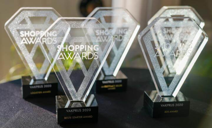 Vijf transparante diamantvormige trofeeën rusten op een zwart oppervlak, elk met het label Shopping Awards 2020. Ze vieren categorieën als Vakprijs 2020 en Beste Starter Award, en laten een aantal van Nederlands snelst groeiende bedrijven zien. Zonlicht werpt reflecties op de trofeeën.