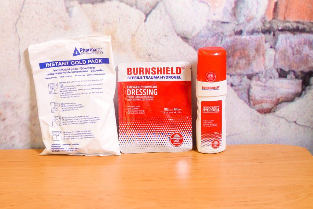 Op een tafelblad staan drie items: een instant coldpack, een Burnshield steriel trauma hydrogelverband en een Burnshield hydrogelspray - essentials voor het snel behandelen van brandwonden (Zo verzorg je snel een brandwond). De achtergrond is een gestructureerd oppervlak dat lijkt op steen of baksteen.